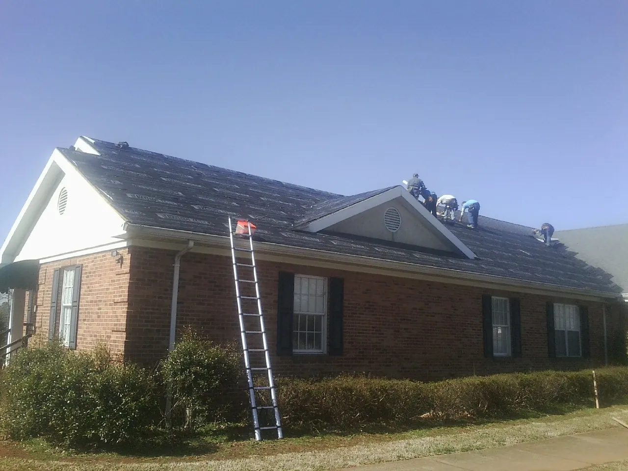 Crew tearing shingles off a brick ranch roof with a ladder set — Metal Roof Repair work by our crew in Lake Waccamaw