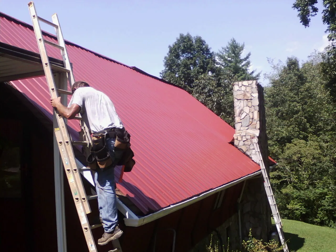 Roofer on a ladder beside a red metal roof and stone chimney — Soffit & Fascia Repair by Roofing Brothers in Lake Waccamaw